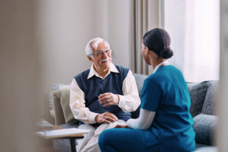 An elderly man sits on a sofa, having a heartfelt conversation with a caring healthcare worker. The scene depicts warmth, trust, and professional caregiving in a comfortable home environment.